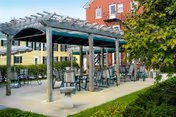Outdoor seating area with multiple tables and chairs under a wooden pergola with green fabric covering. The area is paved and surrounded by greenery, with a red brick building and a yellow building visible in the background.