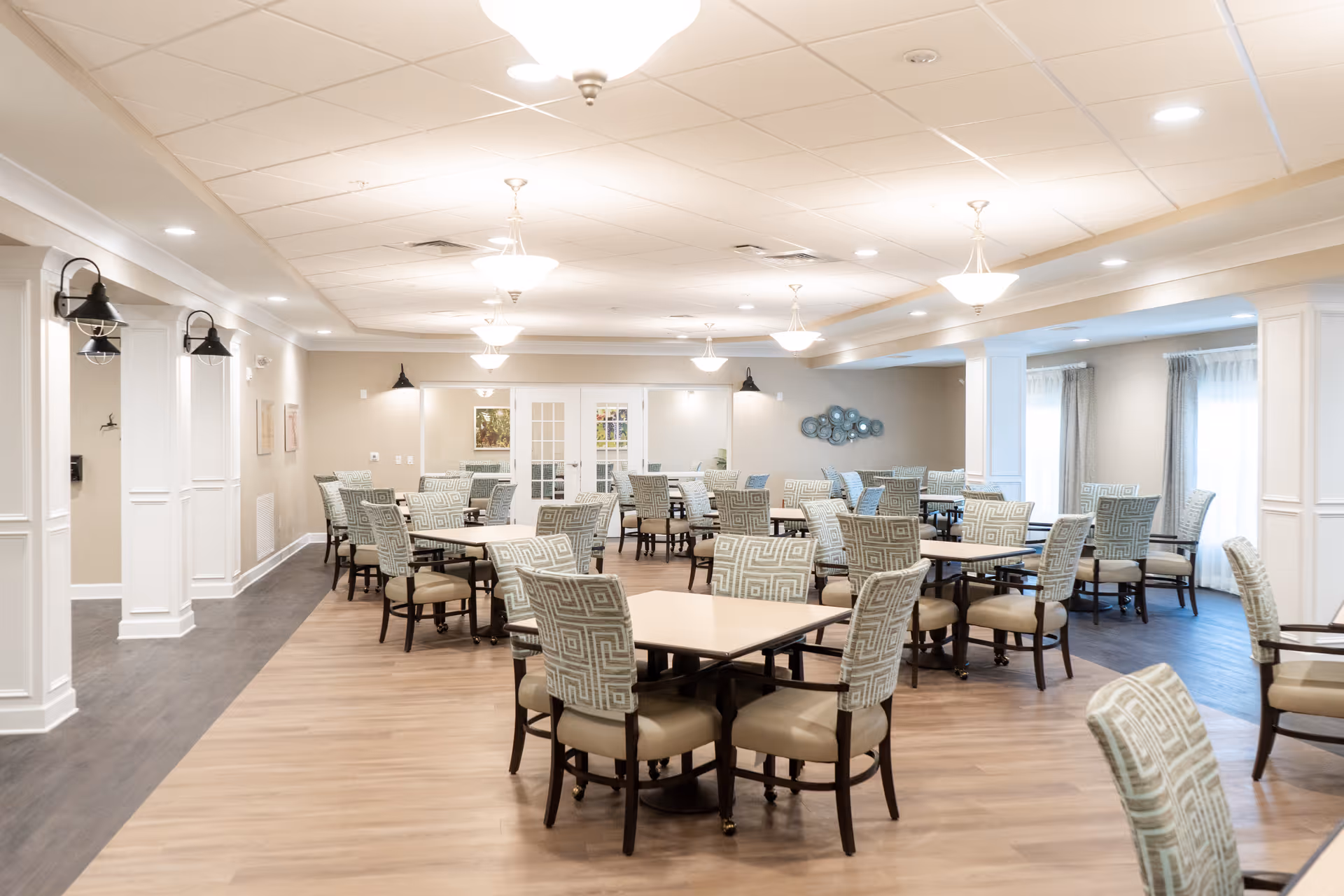 A spacious and well-lit dining room with multiple square tables surrounded by cushioned chairs featuring a geometric pattern. The room has light-colored walls, ceiling lights, and large windows with curtains allowing natural light to enter. The floor is a combination of wood and darker material, and there are decorative wall sconces and artwork on the walls.