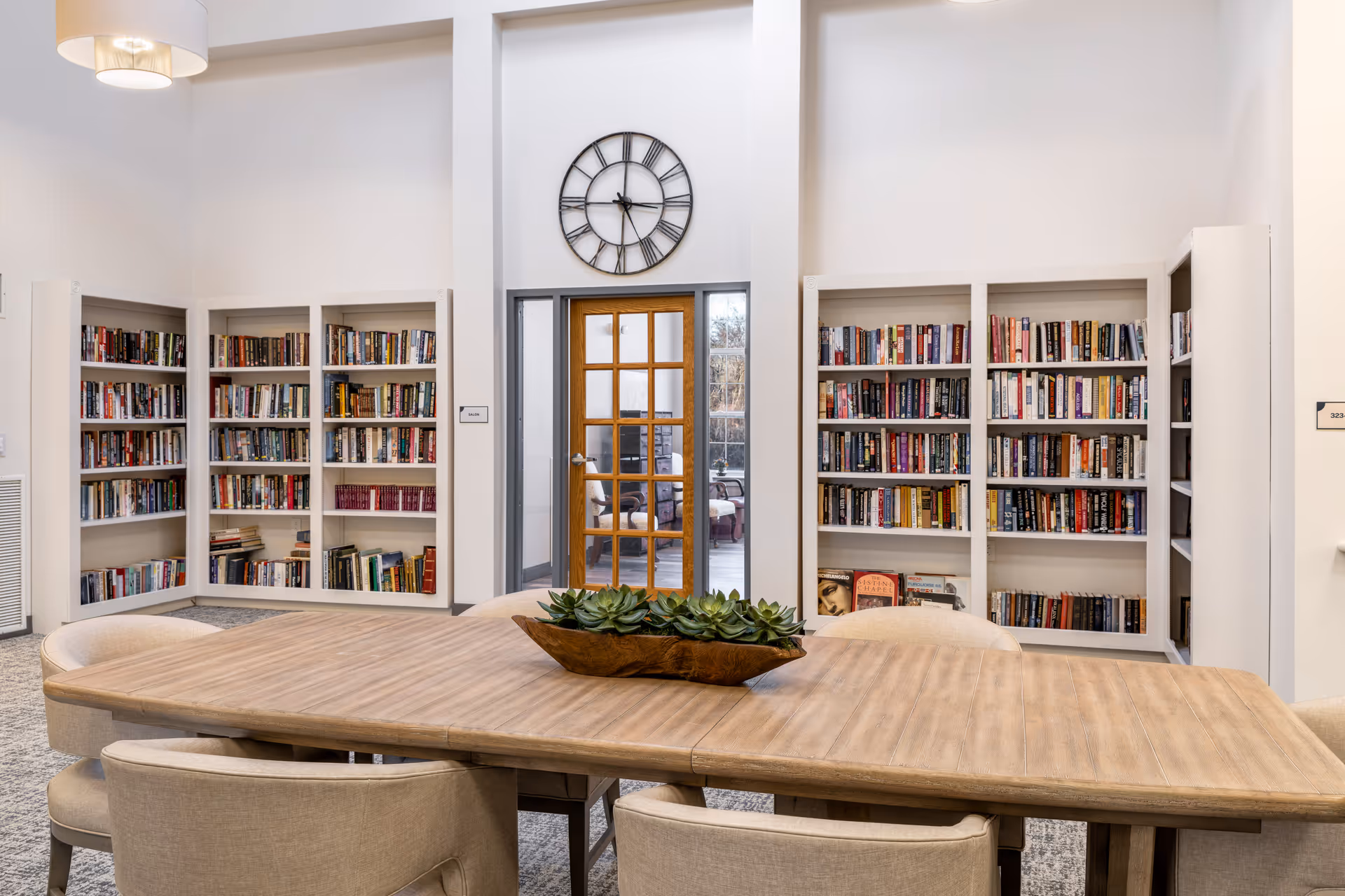 A bright and spacious reading room with built-in bookshelves filled with books along the walls. A large wooden table with beige upholstered chairs surrounds it, and a decorative planter with green succulents sits on the table. A wooden door with glass panels is centered on the far wall beneath a large round wall clock.