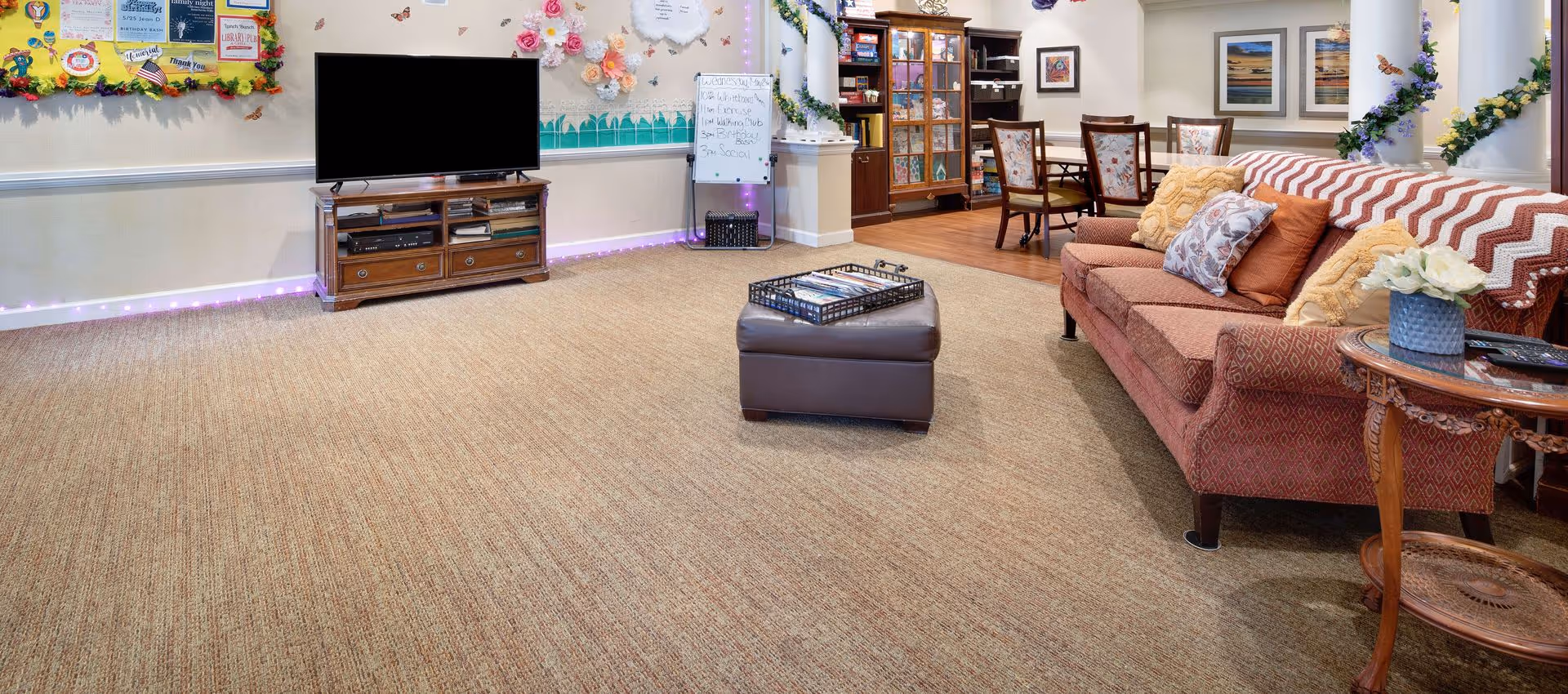 Communal lounge with a patterned sofa, ottoman, flat-screen TV, and a dining table in a decorated senior living common area.