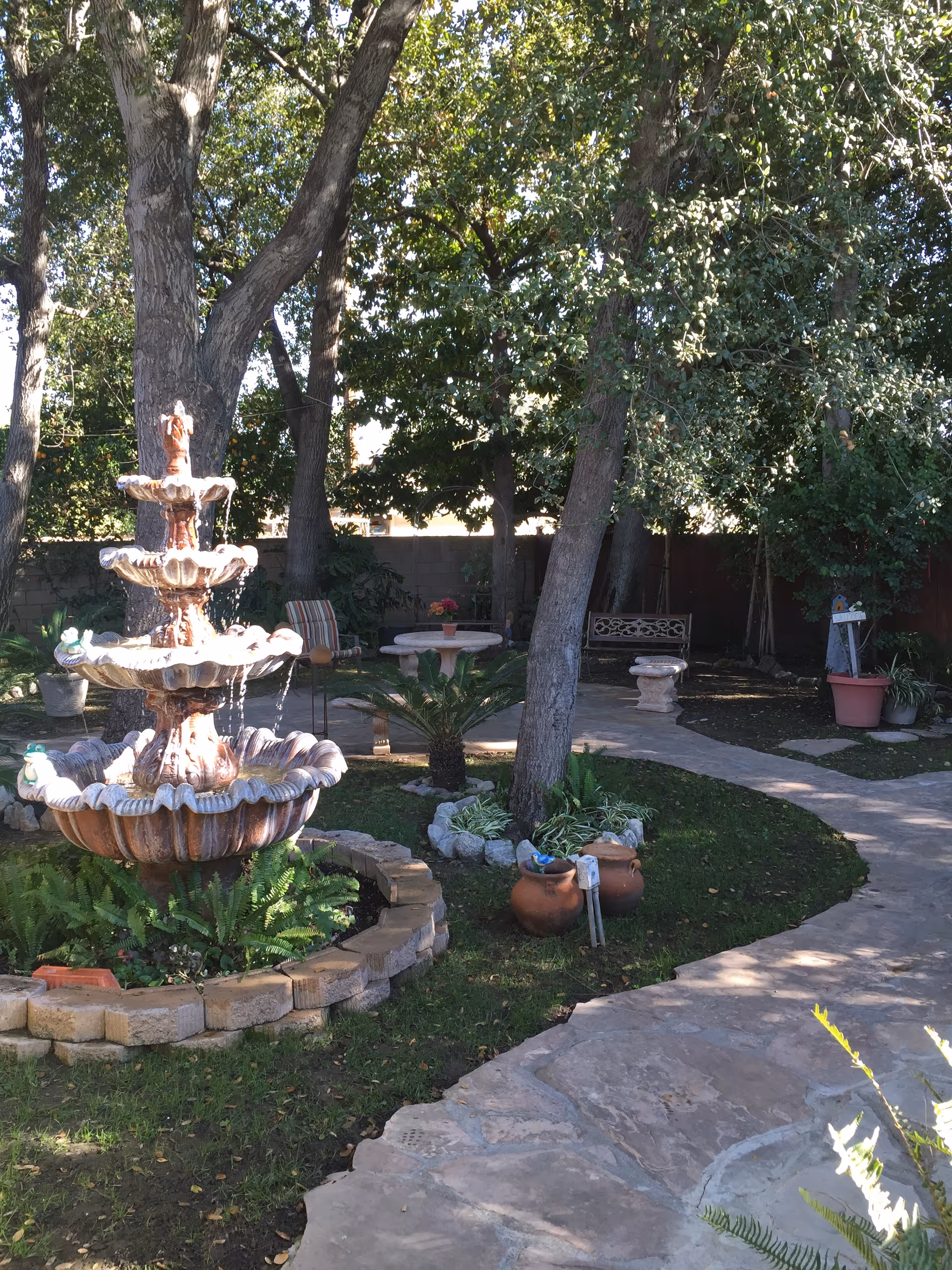 A peaceful outdoor garden area with a multi-tiered stone fountain surrounded by greenery and trees. There are stone pathways, potted plants, a bench, and a round table with chairs in the shaded background.