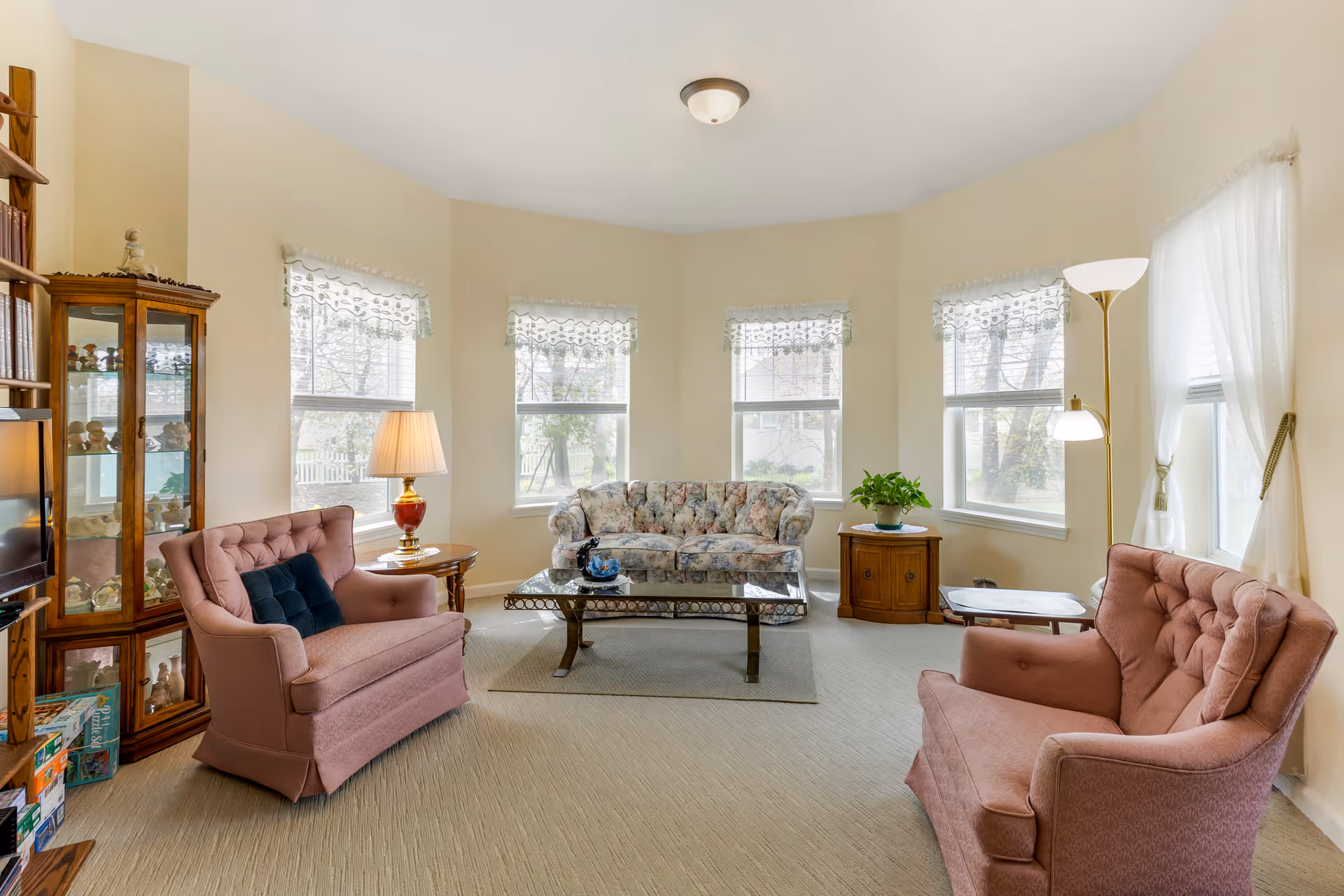 A bright living room with beige walls and carpet, featuring a floral-patterned sofa, two pink upholstered armchairs, a glass coffee table, a wooden side table with a lamp, a corner cabinet with decorative items, and several windows with white lace curtains letting in natural light.