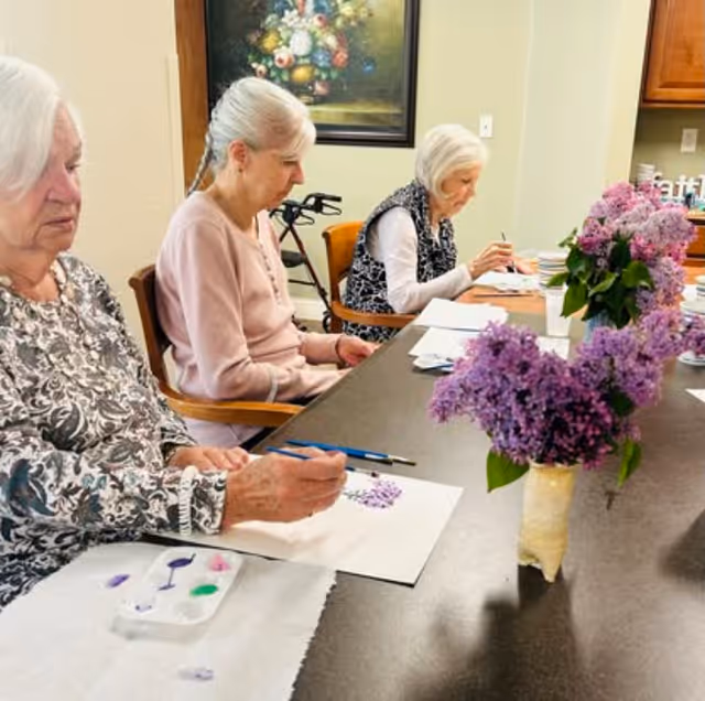 Three elderly women seated at a table engaged in painting with watercolors. A vase with purple lilac flowers is placed on the table. The room has light-colored walls, a painting on the wall, and wooden cabinetry in the background.