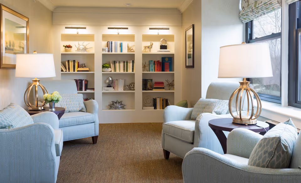 A bright lounge with light-blue upholstered armchairs, side tables with lamps, and built-in bookshelves under wall lighting.