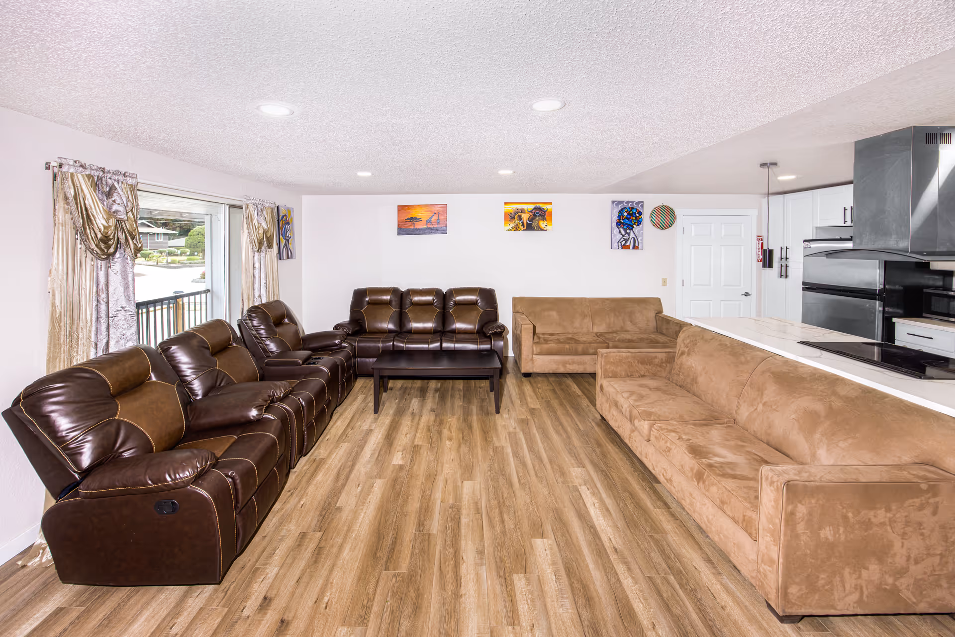 A spacious living room with wood flooring, featuring two brown leather recliner sofas on the left side, a brown fabric sofa on the right, and another brown fabric loveseat against the back wall. The room has white walls adorned with colorful artwork and a large window with decorative curtains on the left. The kitchen area with white cabinets and stainless steel appliances is visible on the right side.