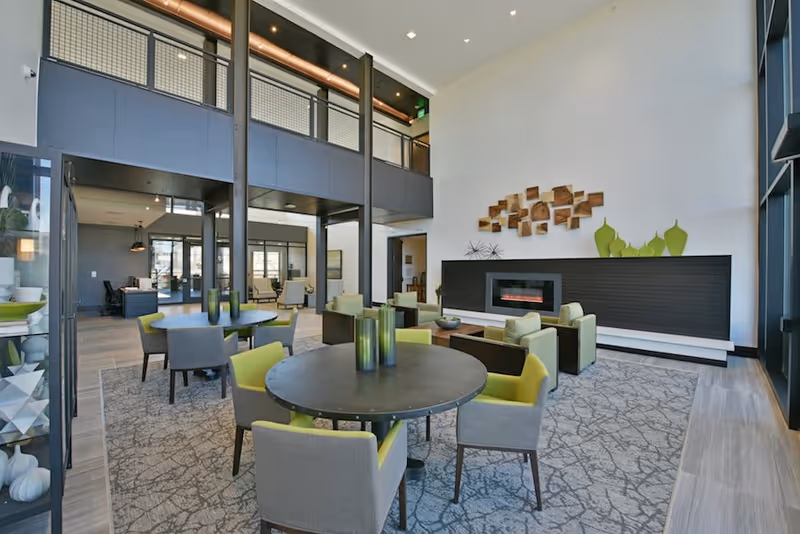 A modern senior living facility common area with round tables surrounded by chairs, a seating area with armchairs near a contemporary electric fireplace, decorative wall art, and large windows allowing natural light.