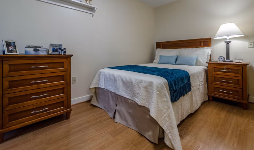 A cozy bedroom with a wooden bed frame, white bedding, and a teal throw blanket. There are two light blue pillows and one teal pillow on the bed. To the right of the bed is a wooden nightstand with a lamp, an alarm clock, and a cup. To the left is a wooden dresser with framed photos and small decorative items on top. The room has light-colored walls and wood flooring.