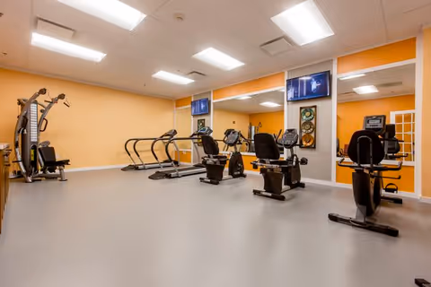 A senior living facility exercise room with various fitness equipment including treadmills, stationary bikes, and a multi-function weight machine. The room has light orange walls, a gray floor, large mirrors on one wall, and two mounted televisions.