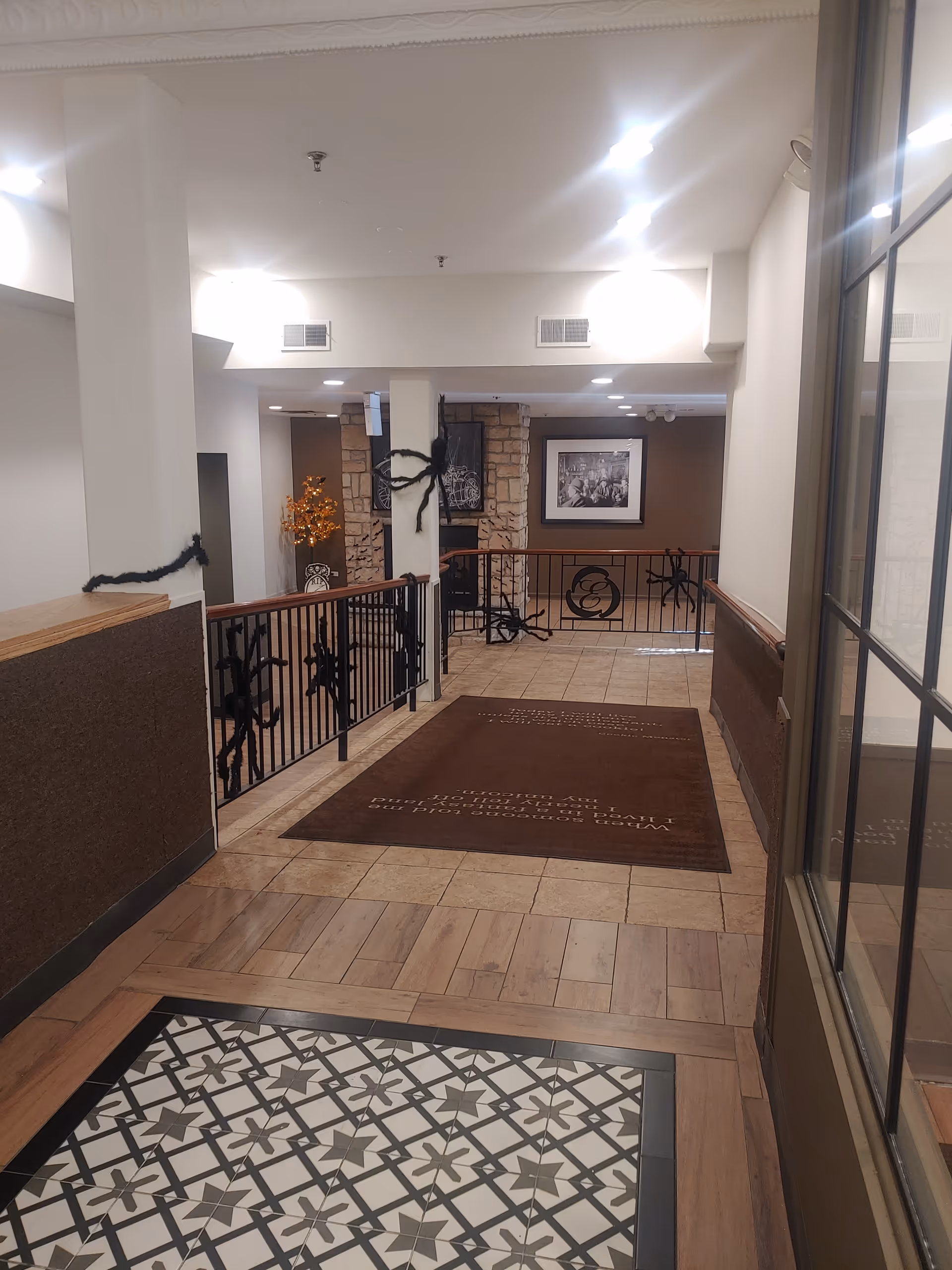 Interior hallway of a senior living facility with tiled and wooden flooring, a brown rug with text, black metal railings decorated with black spider and web decorations, white walls, and ceiling lights. A framed picture and a decorative orange tree are visible in the background.