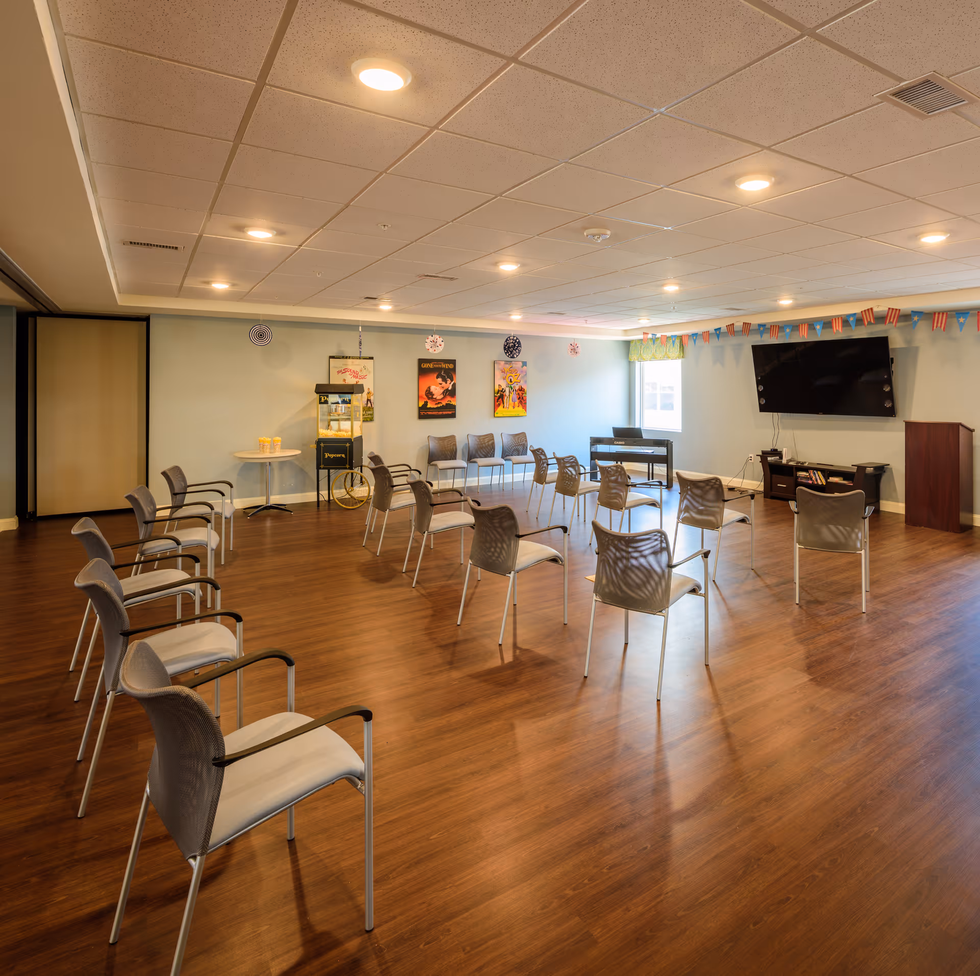A spacious activity room with chairs arranged in a semi-circle on a wooden floor. The room has a popcorn machine, a small table with popcorn containers, a piano, a large flat-screen TV mounted on the wall, and colorful decorations including posters and a string of small flags. The ceiling has recessed lighting and there is a window letting in natural light.