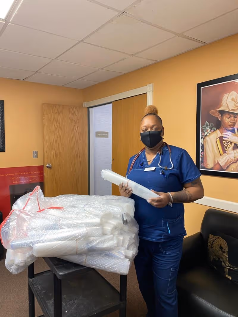 A healthcare worker wearing blue scrubs and a black face mask stands next to a cart filled with wrapped items in a room with beige walls. There is a closed wooden door and a framed painting of a person wearing a hat on the wall behind her.