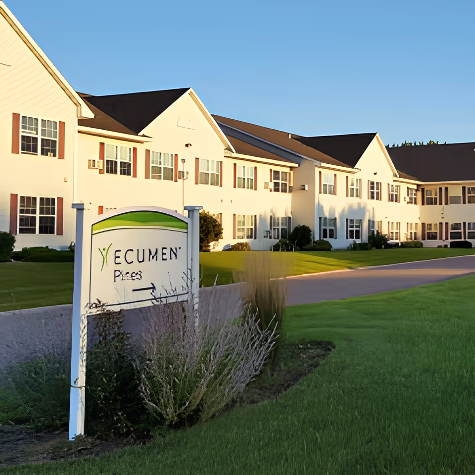 Exterior view of a large, two-story senior living facility building with multiple windows and a dark roof under a clear blue sky. In the foreground, there is a sign that reads 'Ecumen Pines' with some landscaping around it, including grass and bushes.