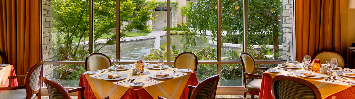 Dining room with round tables set for a meal beside large windows overlooking a pond and garden.