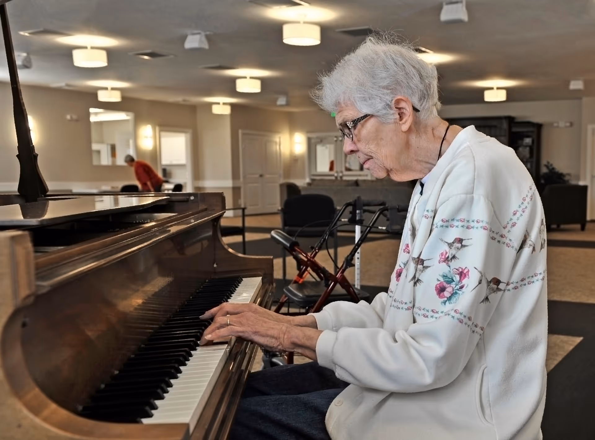 An elderly woman with white hair and glasses is playing a piano in a spacious, well-lit common area of a senior living facility. She is wearing a white sweater with floral and bird patterns. In the background, another person is visible, and there are chairs, tables, and soft lighting fixtures on the ceiling.