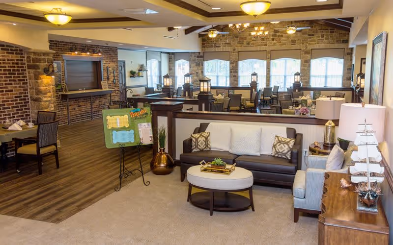 A cozy and well-lit common area in a senior living facility featuring a brown leather sofa with decorative pillows, a round ottoman, an armchair, and a wooden side table with a lamp and a decorative ship model. The background shows a dining area with multiple tables and chairs, brick and stone walls, wooden flooring, and large windows letting in natural light.