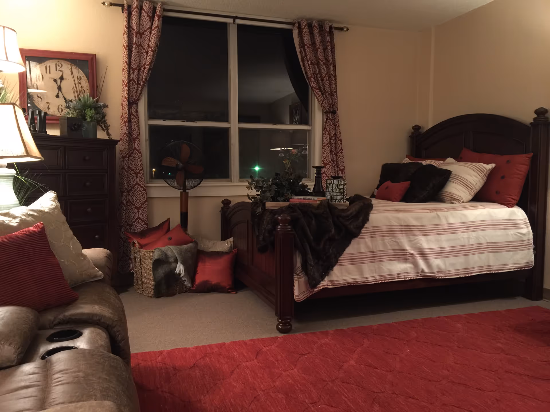 Cozy bedroom with a wooden bed frame, striped bedding, multiple pillows, a dark throw blanket, a wooden dresser with a large clock and plants on top, a basket with pillows and a blanket, a window with patterned red curtains, a floor fan, a beige recliner sofa with red and cream pillows, and a red carpet on the floor.