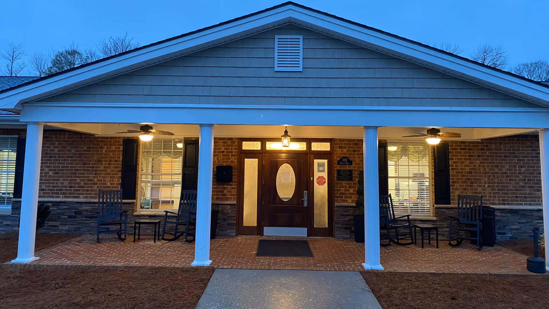 Front entrance of a single-story brick building with a covered porch supported by white columns. The porch has two ceiling fans with lights, four rocking chairs, and two small tables. The entrance door is wooden with glass panels and a light above it. The ground is covered with red bricks and a concrete walkway leads to the door.