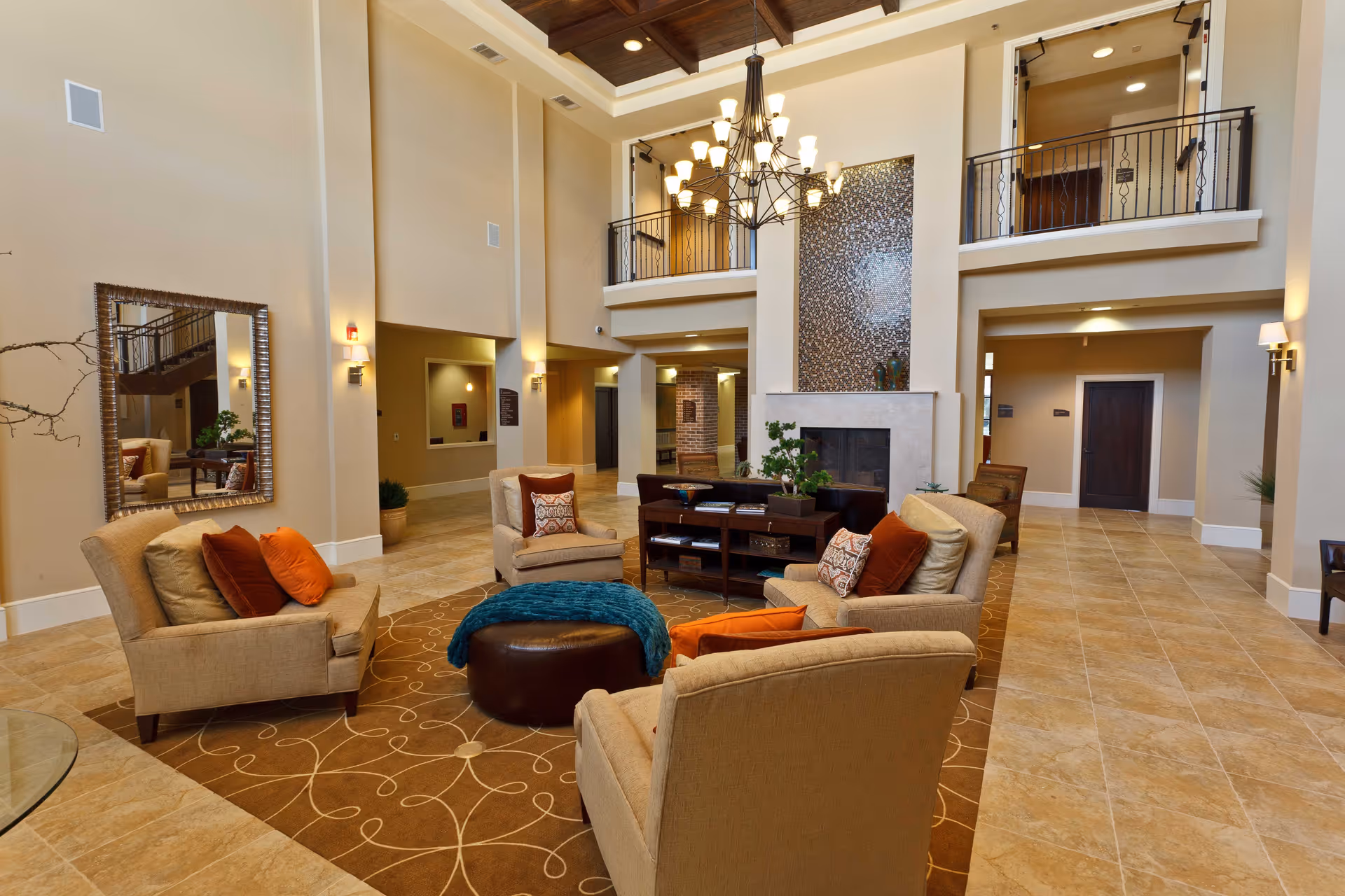 Spacious and elegant living room area with high ceilings, a large chandelier, beige walls, and tiled floors. The room features comfortable beige armchairs with orange and patterned cushions arranged around a brown ottoman and a dark wood coffee table. A large mirror is mounted on one wall, and there is a fireplace with decorative tiles above it. The room has an open balcony on the upper level with black railings.