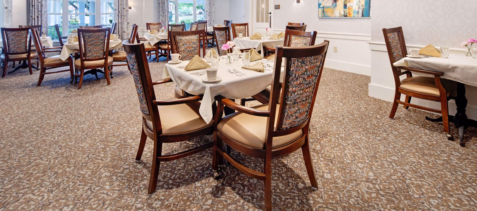 A bright dining room with multiple tables covered in white tablecloths, each set with beige napkins, cups, glasses, and silverware. The chairs have wooden frames with patterned fabric backs and beige cushioned seats. Large windows with curtains allow natural light to fill the room, and a painting hangs on the wall.