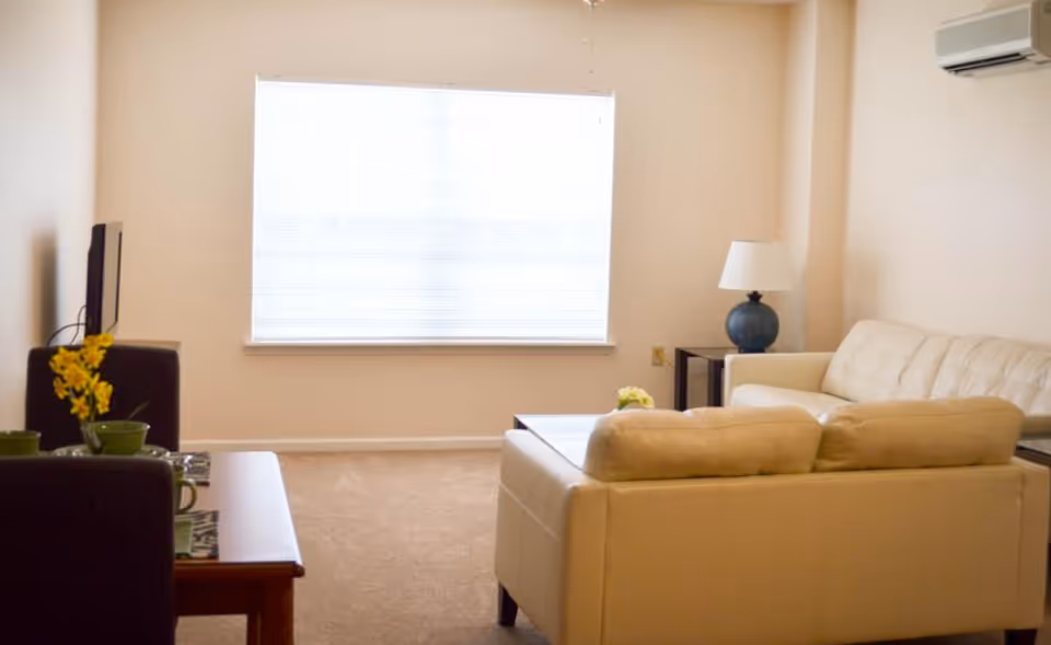 Bright living room with cream leather sofas, a coffee table, a TV, and a large window with blinds.