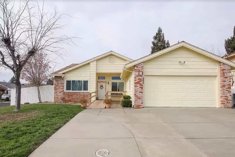Front exterior view of a single-story house with a two-car garage, a concrete driveway, a small porch with potted plants, and a leafless tree on the left side of the lawn.