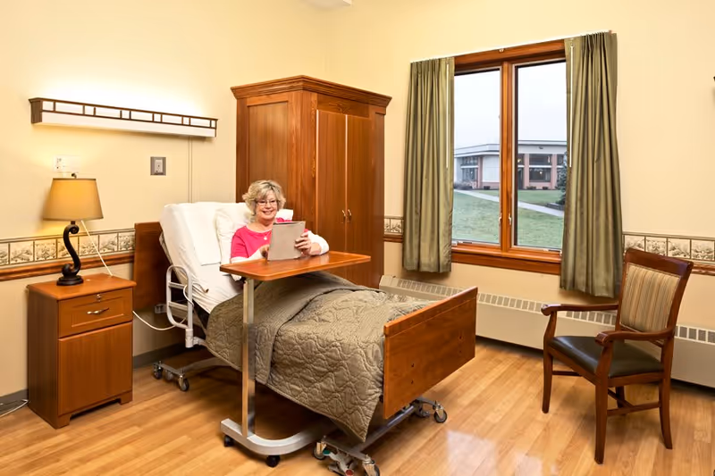 A senior woman sitting up in a hospital-style bed in a well-lit room, using a tablet. The room has wooden furniture including a nightstand with a lamp, a wardrobe, and a chair. There is a window with green curtains showing an outside view of a building and grassy area.