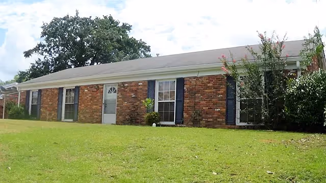 Single-story brick residential building with a gray shingled roof, several windows with dark shutters, a white door, and a grassy lawn in front. Trees and bushes are visible around the building under a partly cloudy sky.