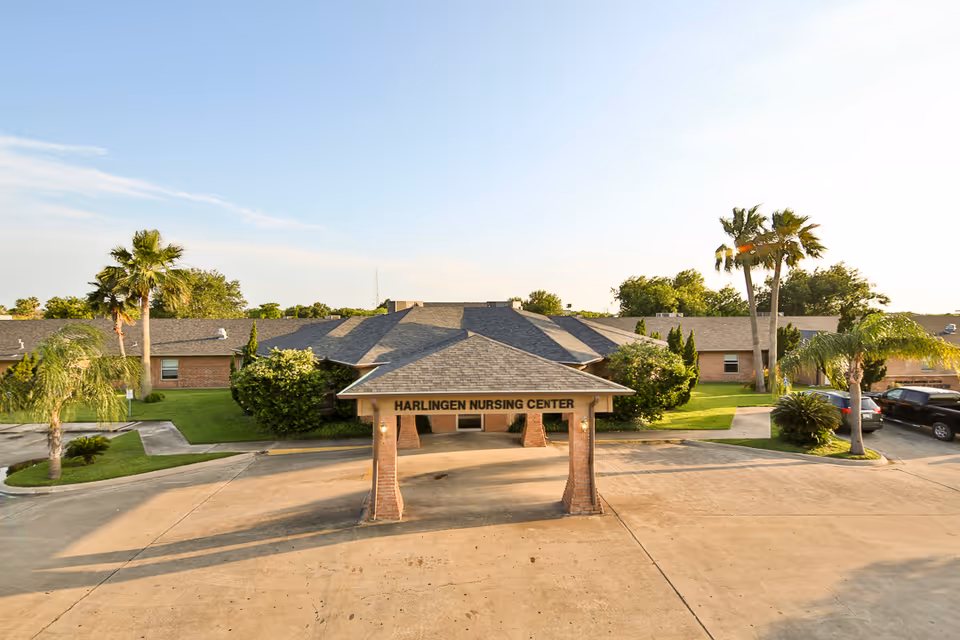 Exterior view of Harlingen Nursing and Rehabilitation Center showing a single-story brick building with a covered entrance supported by brick pillars. The area around the building is landscaped with palm trees, bushes, and green grass under a clear sky.
