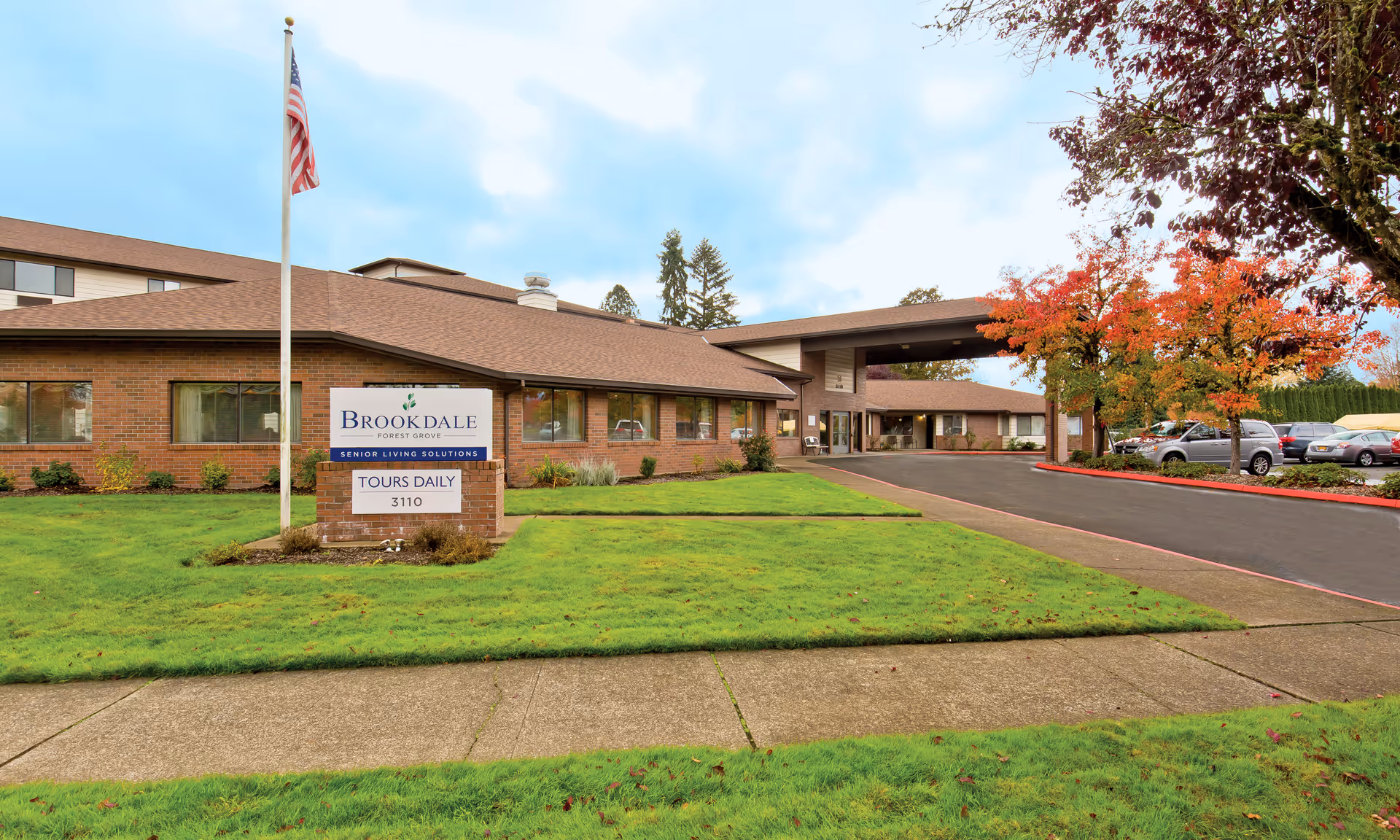 Exterior view of Brookdale Forest Grove senior living facility with a brick building, an American flag on a flagpole, a sign indicating tours daily, a driveway, and a parking lot with cars. Trees with autumn-colored leaves are visible on the right side.