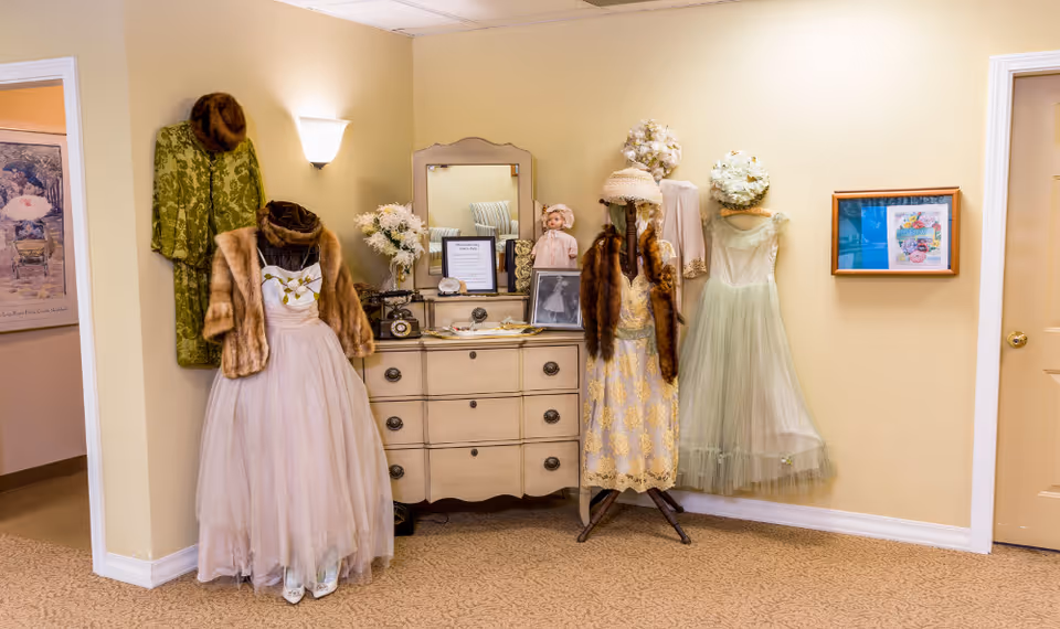 A vintage-style room corner with mannequins dressed in old-fashioned dresses and fur coats, a cream-colored dresser with a mirror, decorative flowers, framed pictures on the wall, and a soft yellow wall color.