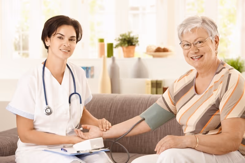 A healthcare professional in a white uniform with a stethoscope around her neck is measuring the blood pressure of a smiling elderly woman sitting on a couch in a bright, well-lit room with decorative vases and plants in the background.