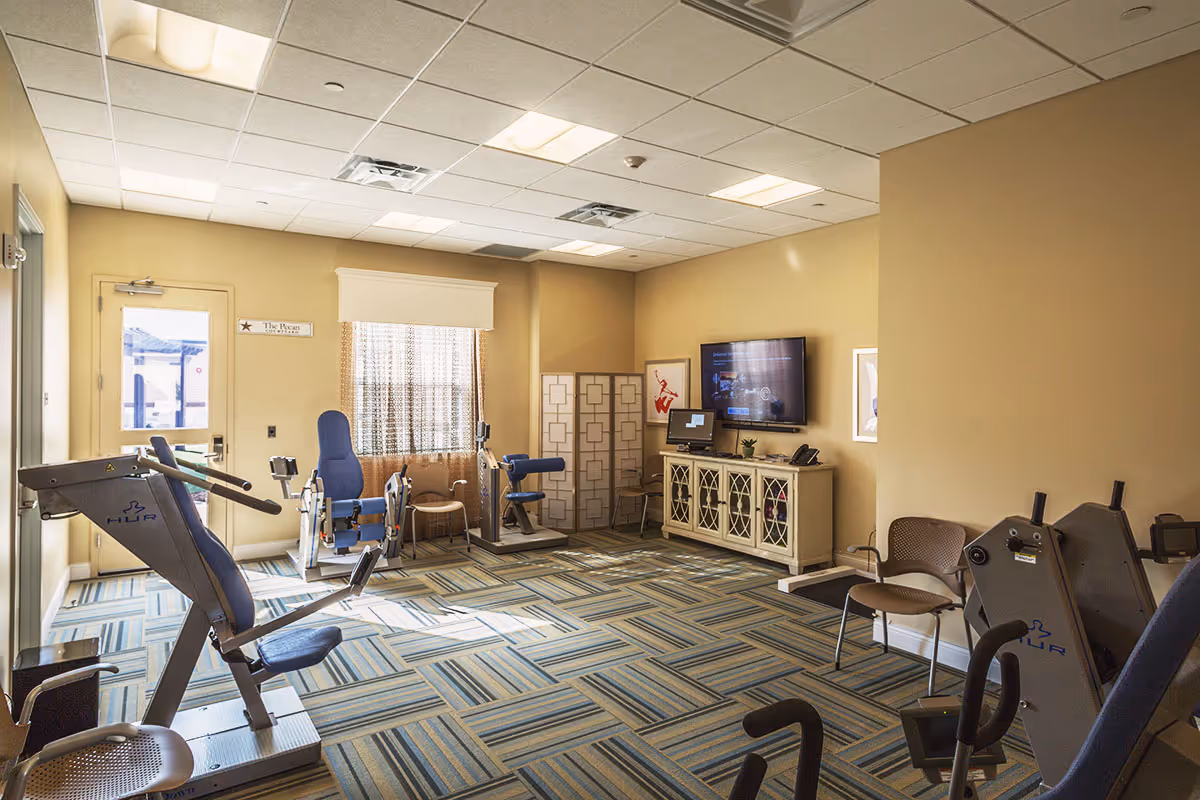 Well-lit interior exercise room with multiple pieces of fitness equipment, chairs, a wall-mounted TV, and patterned carpet.