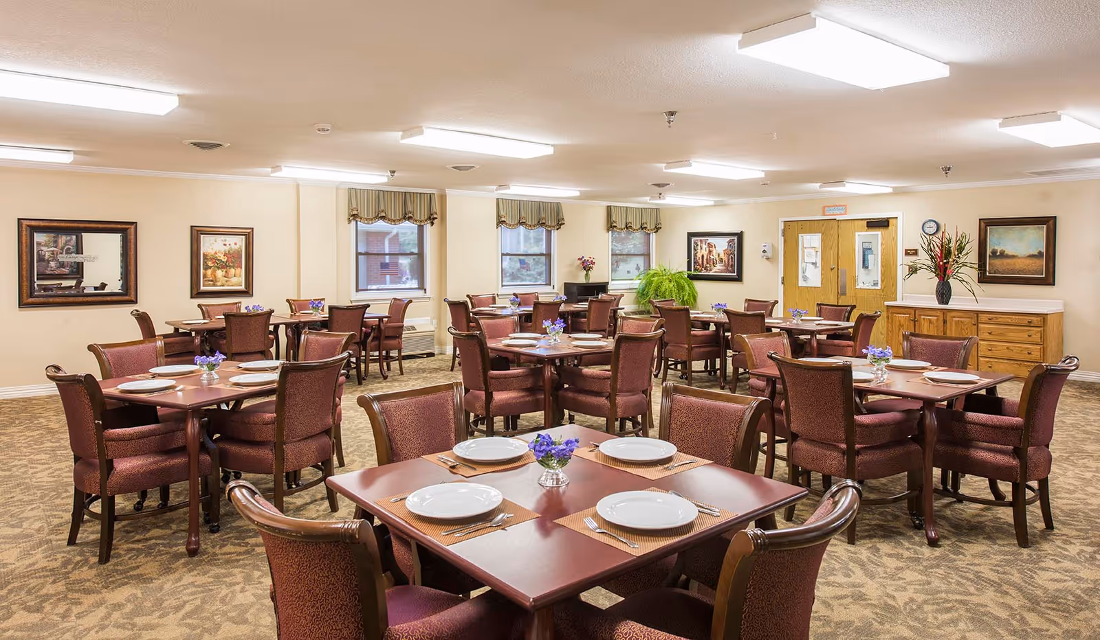 Spacious dining room in a senior living facility with multiple set tables, upholstered chairs, floral centerpieces and warm overhead lighting.