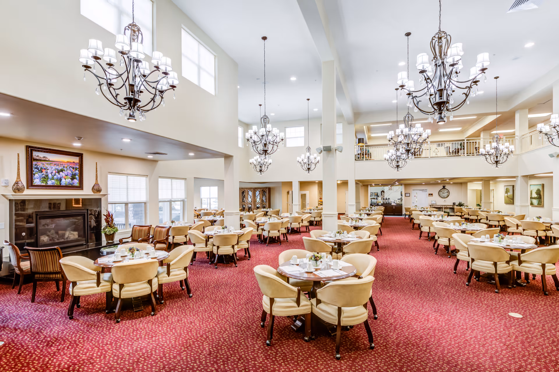 Spacious dining room with multiple round tables surrounded by beige upholstered chairs, red patterned carpet, large chandeliers hanging from a high ceiling, and a fireplace with a framed painting above it. The room has large windows allowing natural light and a balcony area overlooking the dining space.