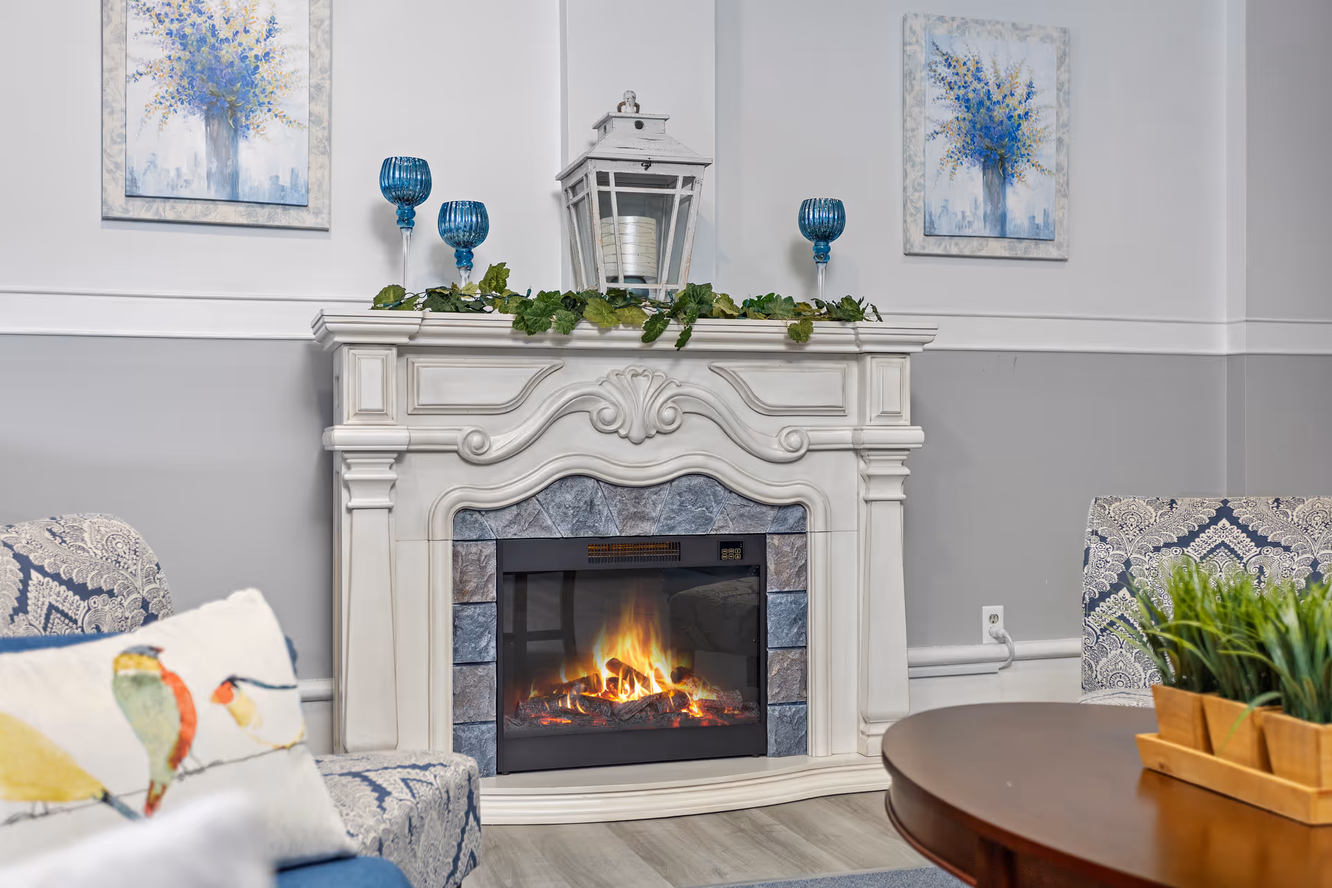 A cozy living room area featuring a white ornate fireplace with a lit electric fire. On top of the mantel are decorative items including a white lantern, green garland, and three blue candle holders. Two framed paintings of blue flowers hang on the wall above the fireplace. Surrounding the fireplace are patterned armchairs and a wooden coffee table with a planter of green plants.