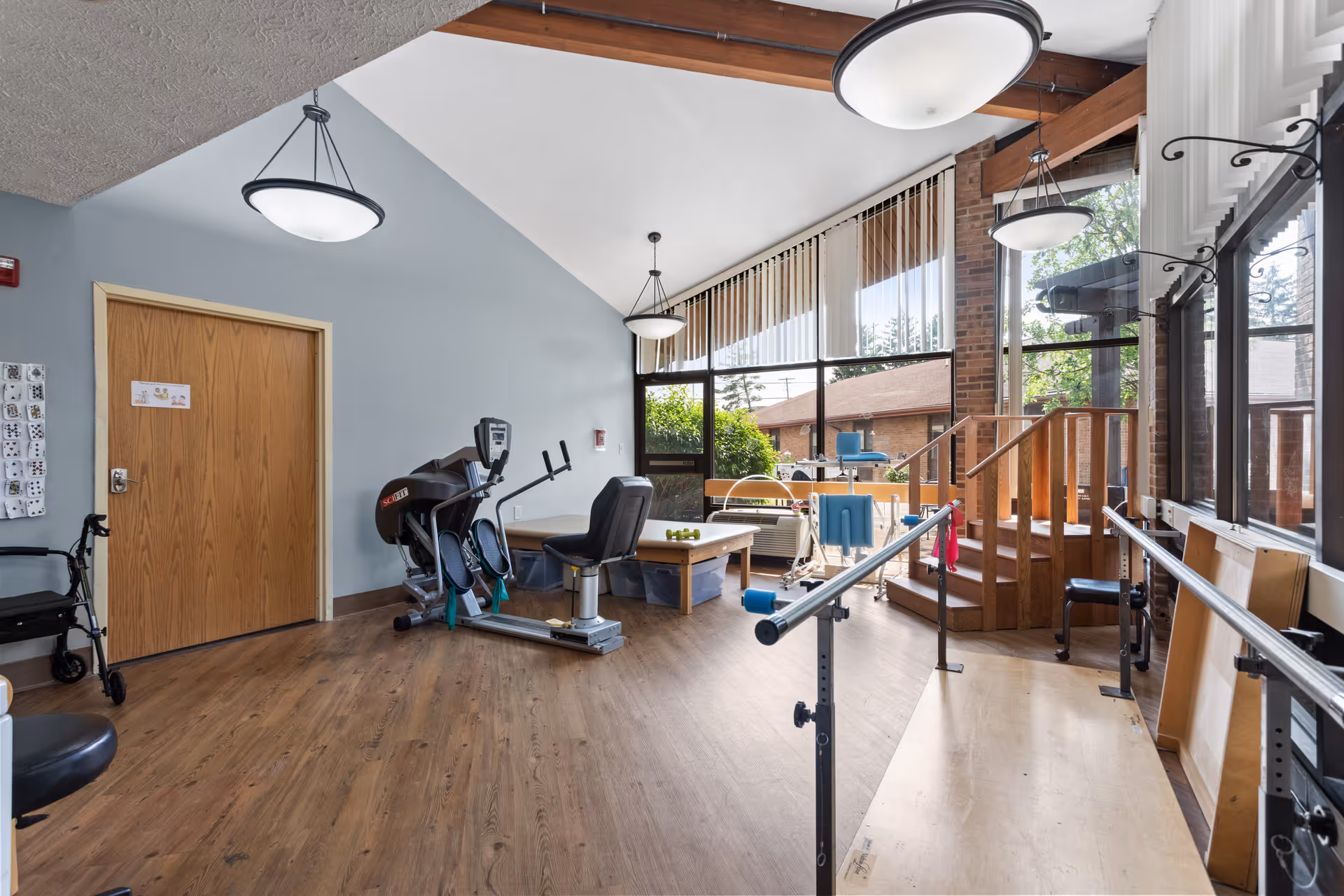 A rehabilitation or physical therapy room with exercise equipment including a stationary bike, parallel bars for walking practice, a walker, and a small set of stairs. The room has large windows letting in natural light, wooden flooring, and modern ceiling lights.