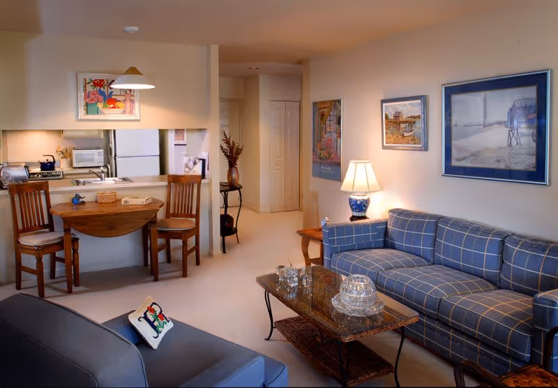 A cozy living room and kitchen area in a senior living facility. The living room features a blue plaid sofa, a glass coffee table with decorative glassware, a side table with a lamp, and framed artwork on the walls. The kitchen area has a small wooden dining table with two chairs, a refrigerator, microwave, and stove. The space is warmly lit and decorated with a vase of flowers on a small table in the hallway.