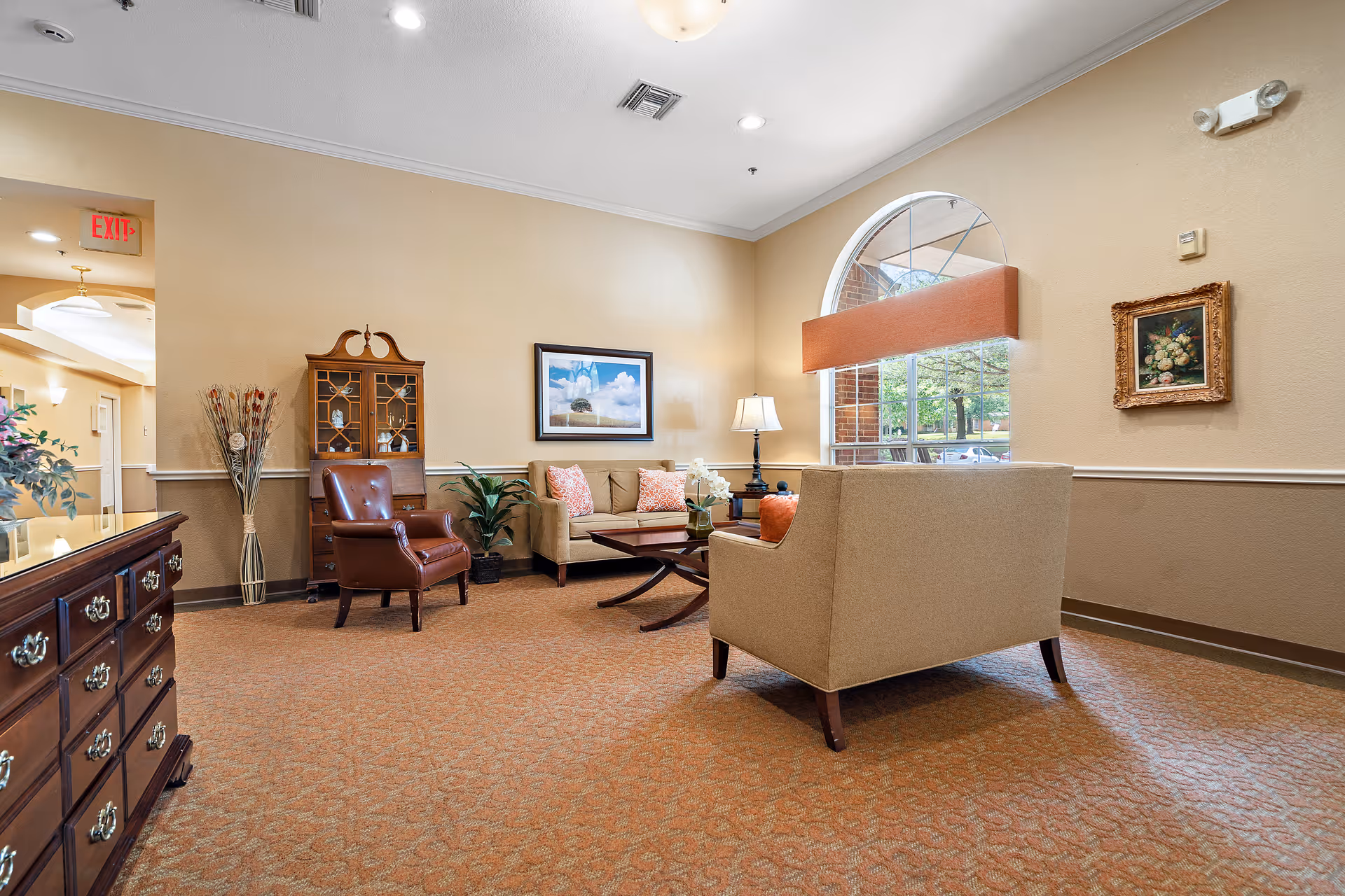 A cozy sitting area in a senior living facility with beige walls and carpet. The room features a beige sofa with patterned pillows, a matching armchair, a brown leather chair, a wooden coffee table, a wooden cabinet with glass doors, a table lamp, and framed artwork on the walls. A large arched window with an orange valance lets in natural light.