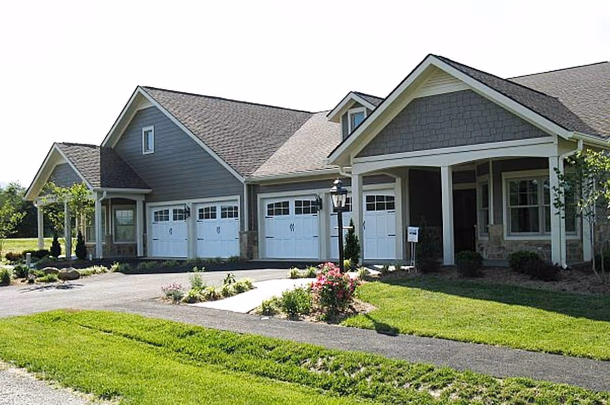 Exterior view of a single-story residential building with multiple garage doors, a covered porch, and well-maintained landscaping including green grass and flowering plants.
