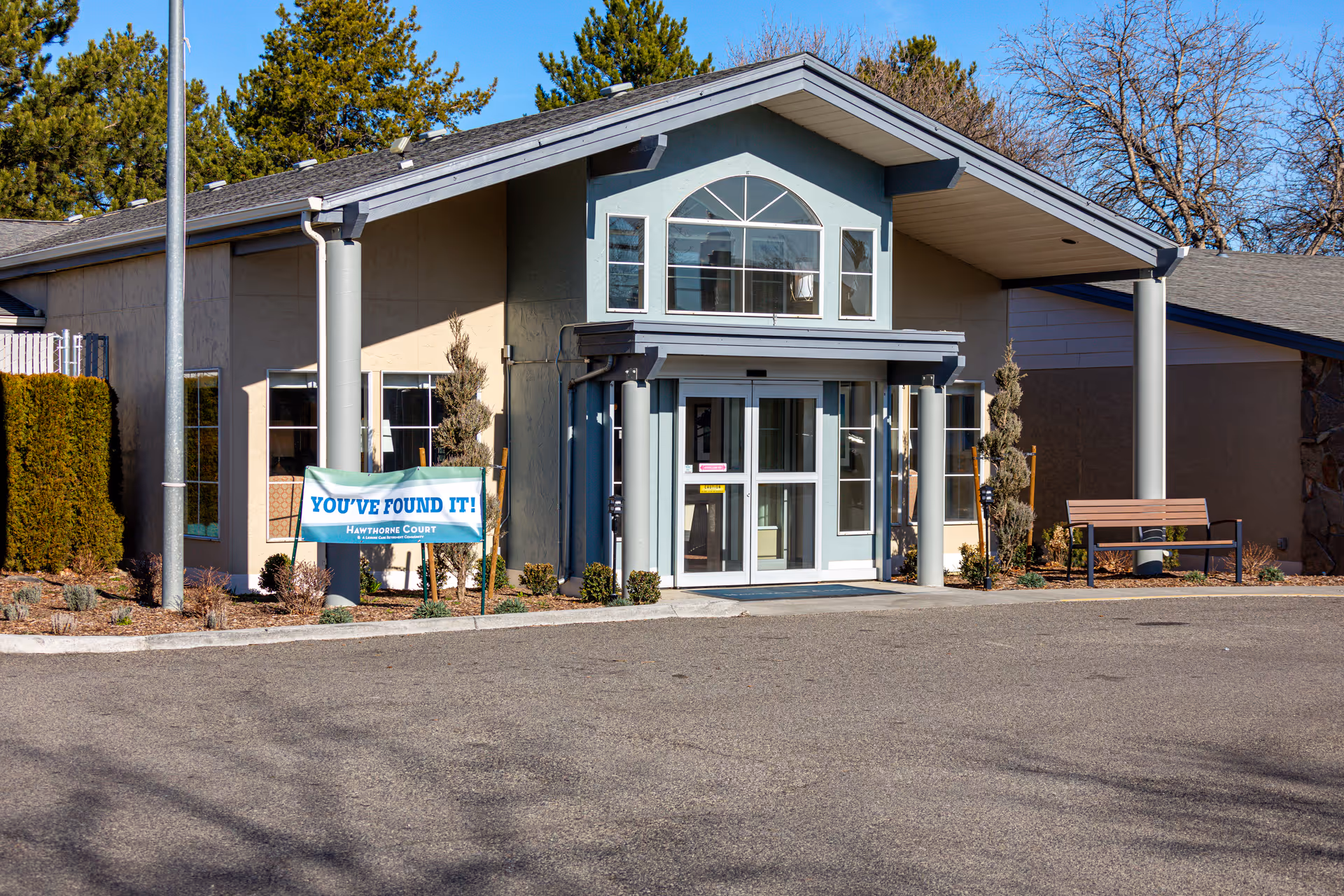 Front entrance of Hawthorne Court with a covered portico, a 'YOU'VE FOUND IT!' banner and a bench nearby.