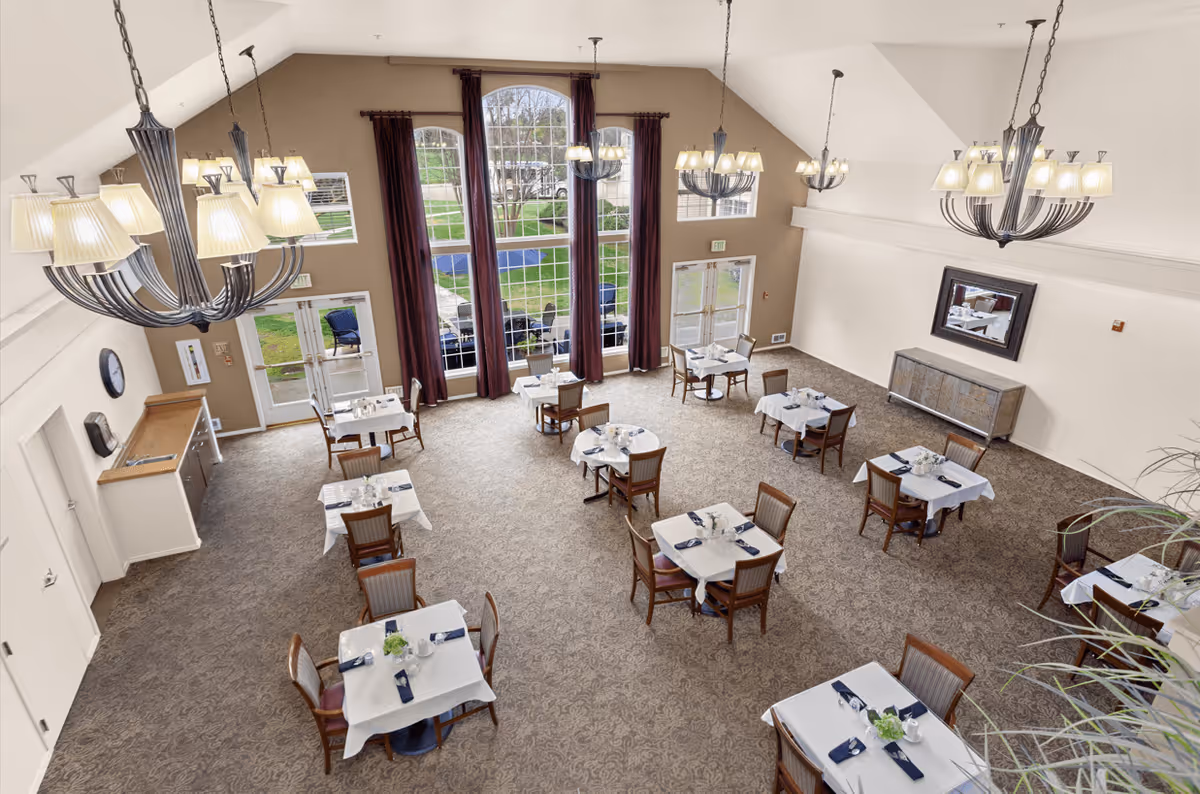 A spacious dining room with multiple square tables covered with white tablecloths, each set with napkins, glasses, and small floral centerpieces. The room has large windows with dark curtains letting in natural light, beige walls, carpeted floor, and several elegant chandeliers hanging from the ceiling.