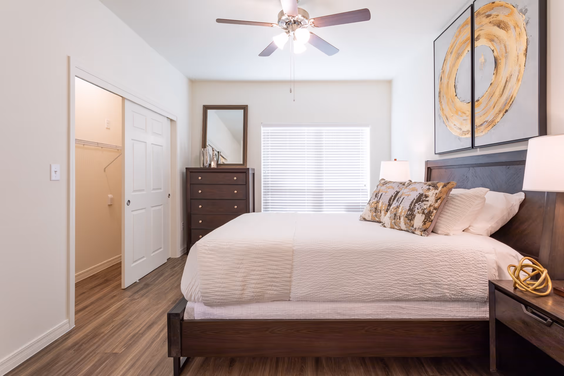 A bright and clean bedroom with a large bed covered in white bedding and decorative pillows. The room features wooden furniture including a dresser with a mirror, a nightstand with a lamp and decorative object, and a ceiling fan with lights. A window with blinds is centered on the far wall, and there is an open closet with sliding doors on the left side.