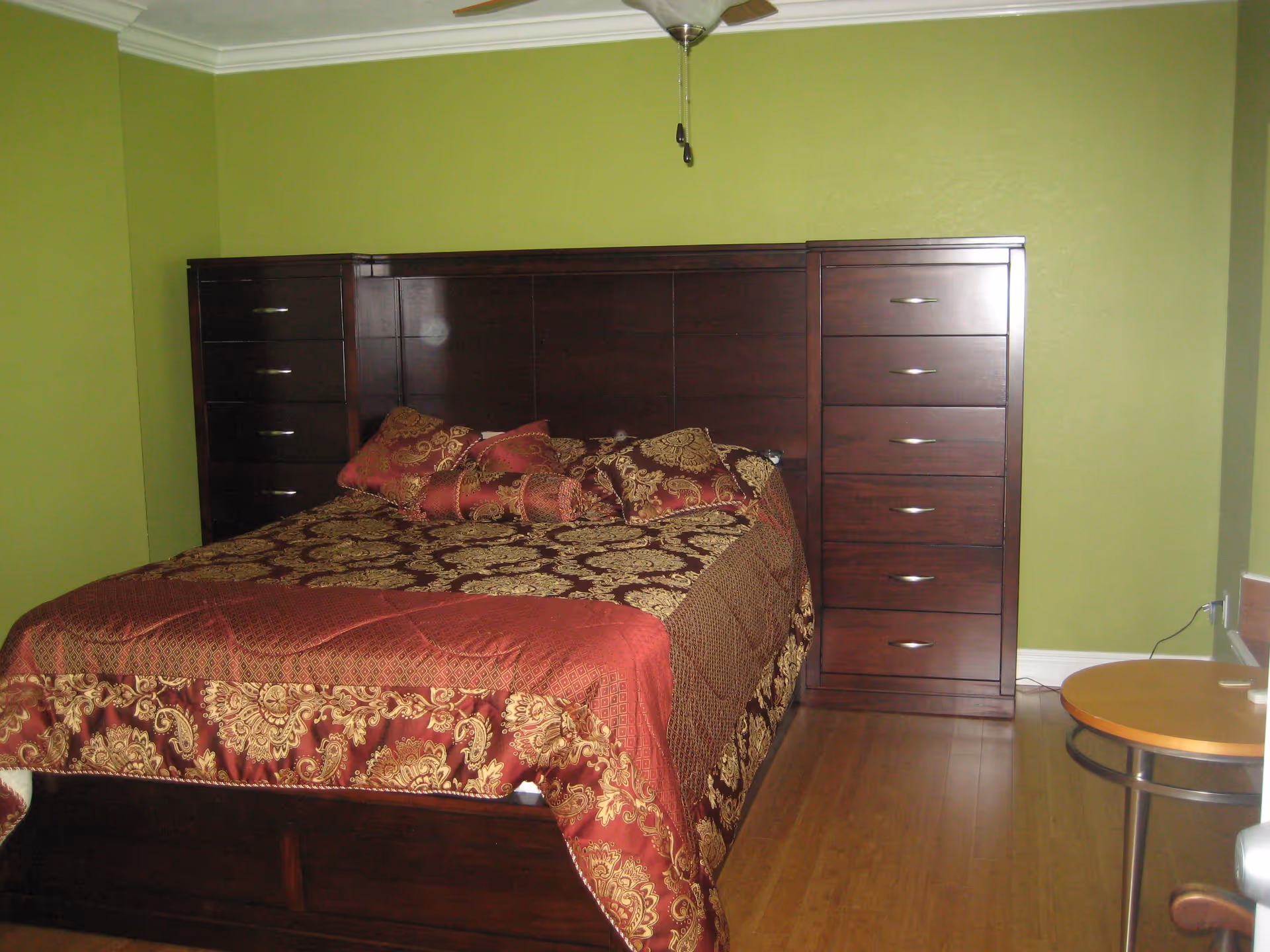 A bedroom with a large dark wooden bed frame featuring built-in drawers on both sides. The bed is covered with a red and gold patterned bedspread and matching pillows. The walls are painted green, and there is a ceiling fan above. A small round wooden table is visible on the right side of the image.
