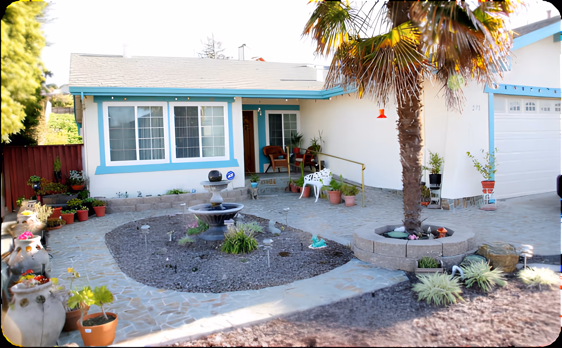 Front yard and entrance of a single-story house with a central fountain, palm tree, potted plants, and turquoise-trimmed windows.