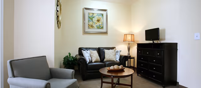 Small living room with a leather loveseat, upholstered armchair, round coffee table, dresser with a TV, lamp, and wall art.