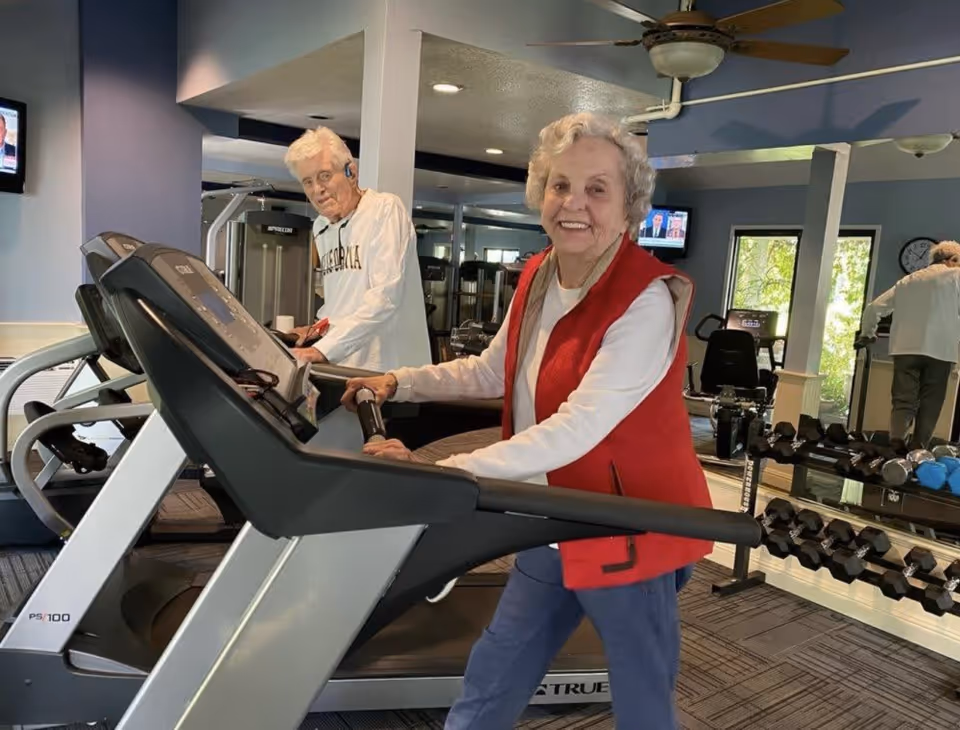 Two elderly individuals exercising on treadmills in a fitness room with mirrors, weights, and exercise equipment. One woman in a red vest is smiling at the camera, while an elderly man in a white sweatshirt is using another treadmill. The room has blue walls, ceiling fans, and a television mounted on the wall.