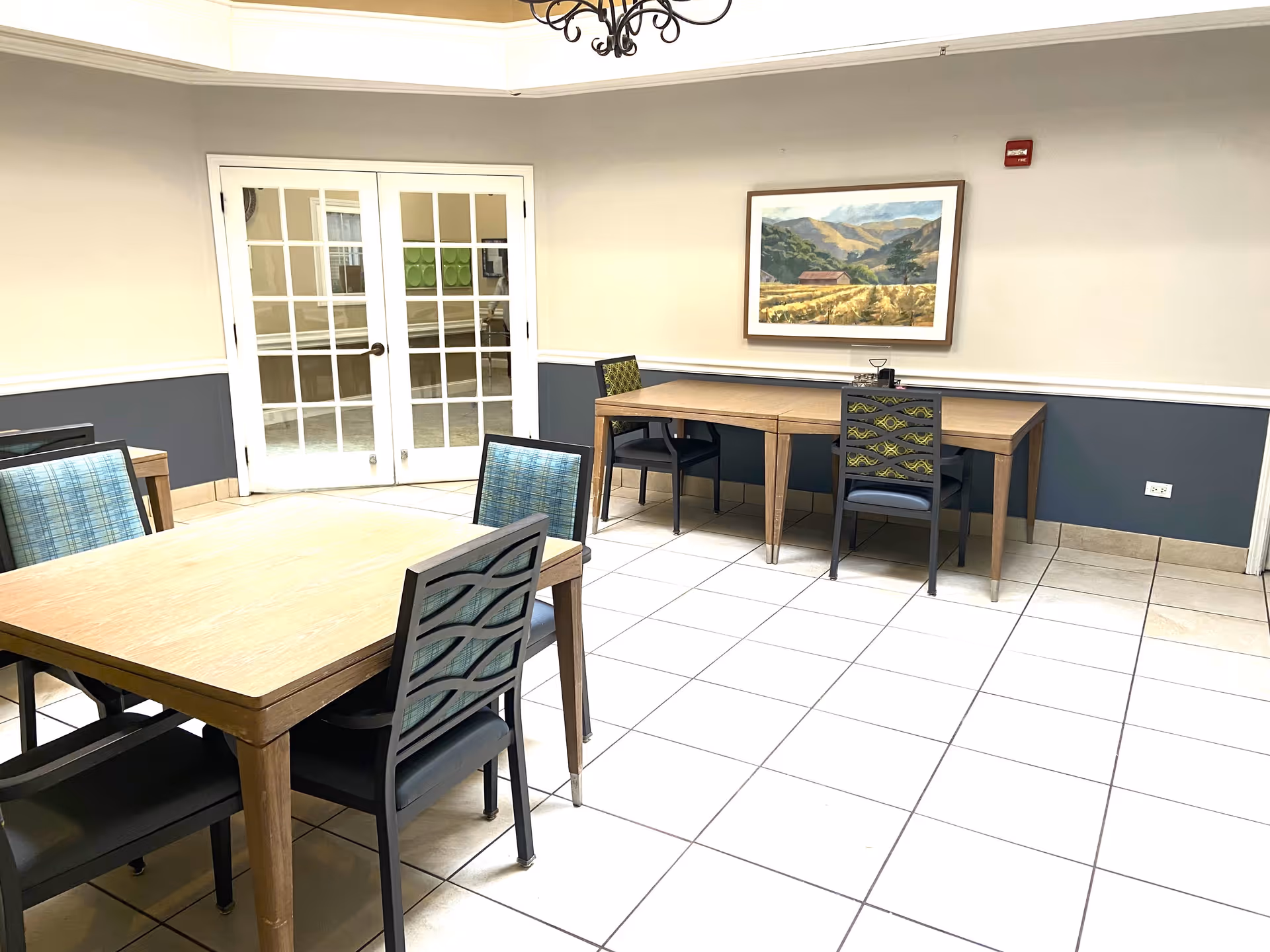 A bright room with tiled floors featuring two wooden tables and several chairs with patterned backs and blue cushions. One table is square with four chairs around it, and the other is rectangular with two chairs. A framed landscape painting hangs on the wall above the rectangular table. Double glass doors with white frames are visible in the background.