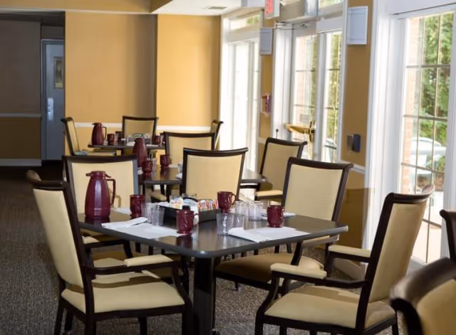 Dining room with tables and chairs set with coffee carafes, mugs, and place settings beside large windows.