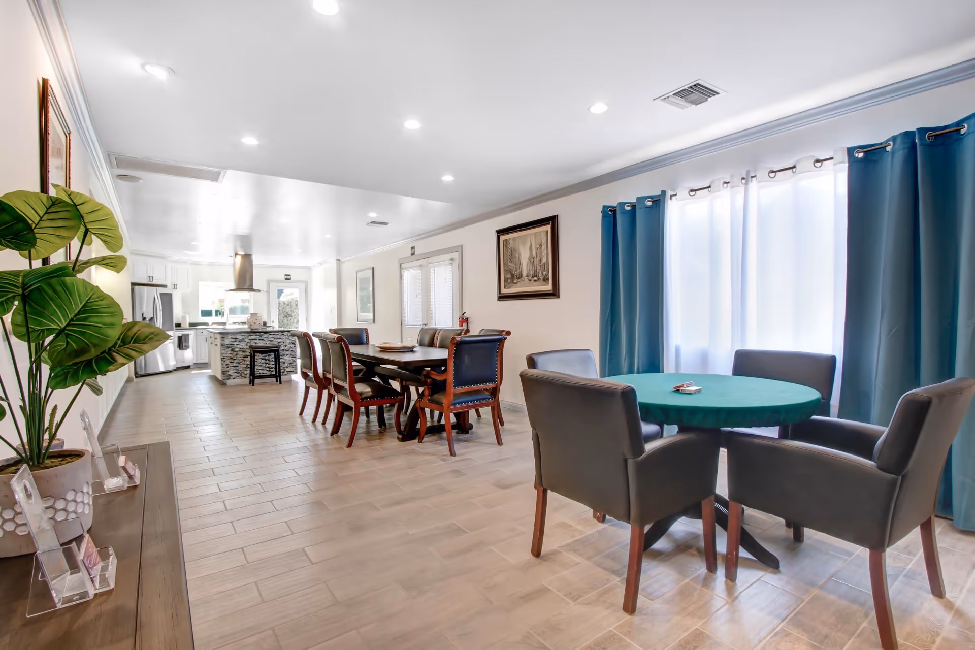 A bright and spacious interior room featuring a round table with four black chairs covered with a green tablecloth near a window with blue curtains. In the background, there is a long dining table with eight chairs and a modern kitchen with stainless steel appliances and an island with a stool. The floor is tiled, and the walls are decorated with framed artwork and a large green potted plant on the left side.