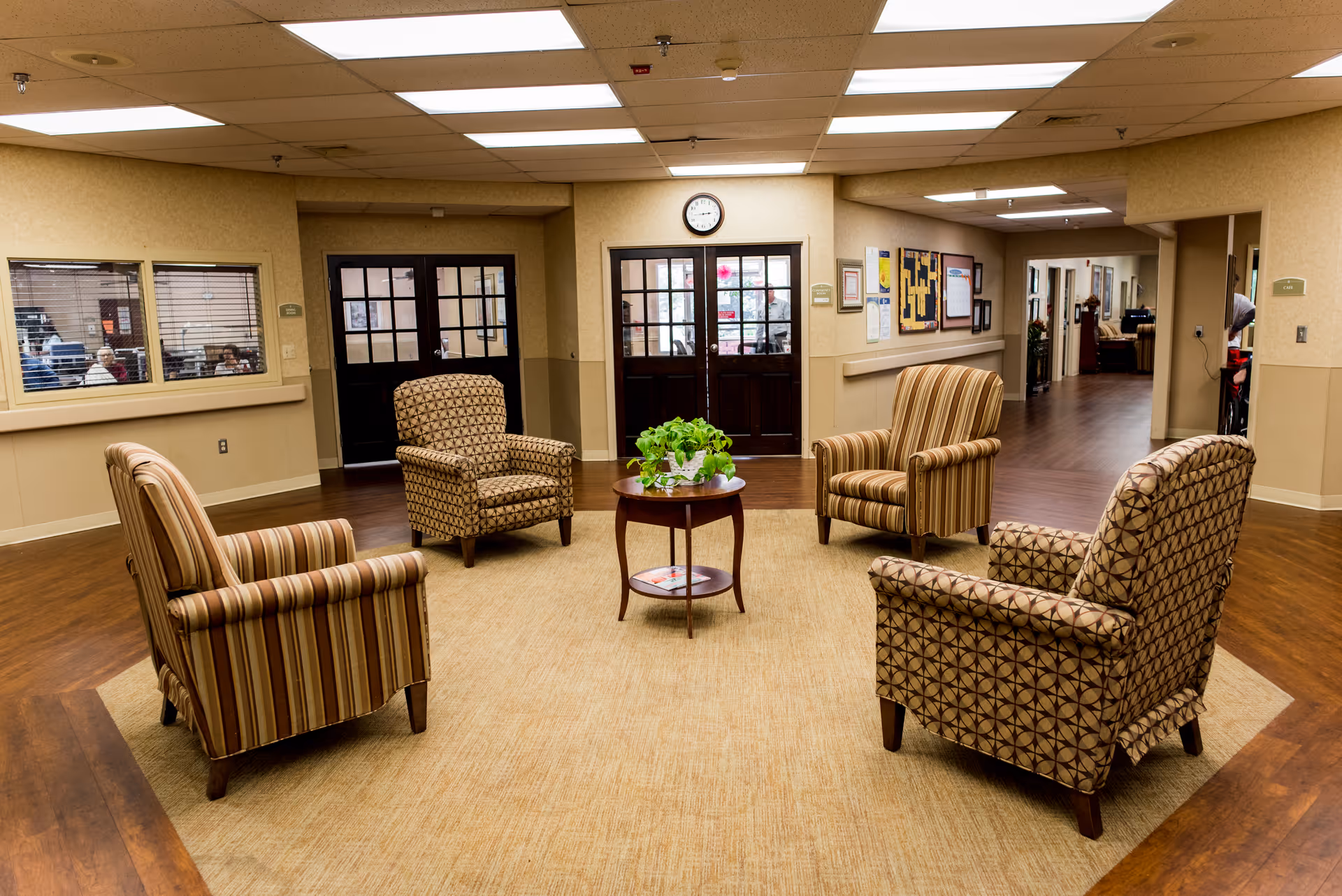 A seating area in a healthcare facility with four patterned armchairs arranged around a small round wooden table with a green plant on it. The room has wood flooring and beige walls, with a hallway and doors visible in the background.